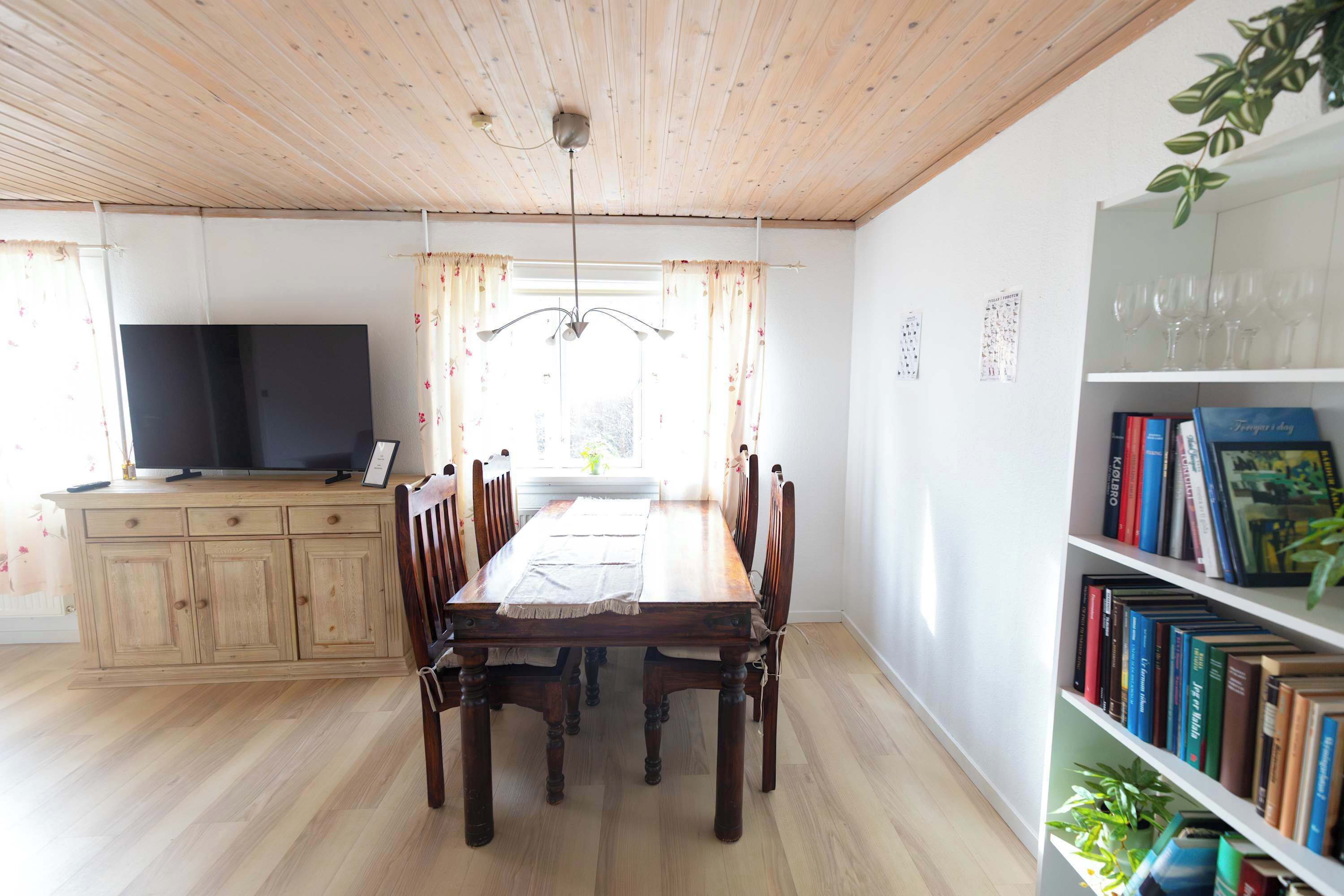 Un comedor con una mesa y sillas de madera, un televisor sobre un mueble bajo, y una estantería llena de libros.