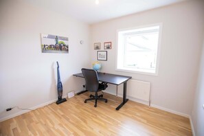 Une pièce dotée d’un plancher en bois, d’un bureau avec une chaise, d’un radiateur, d’une fenêtre et de décorations murales comprenant un tableau et des photos encadrées.