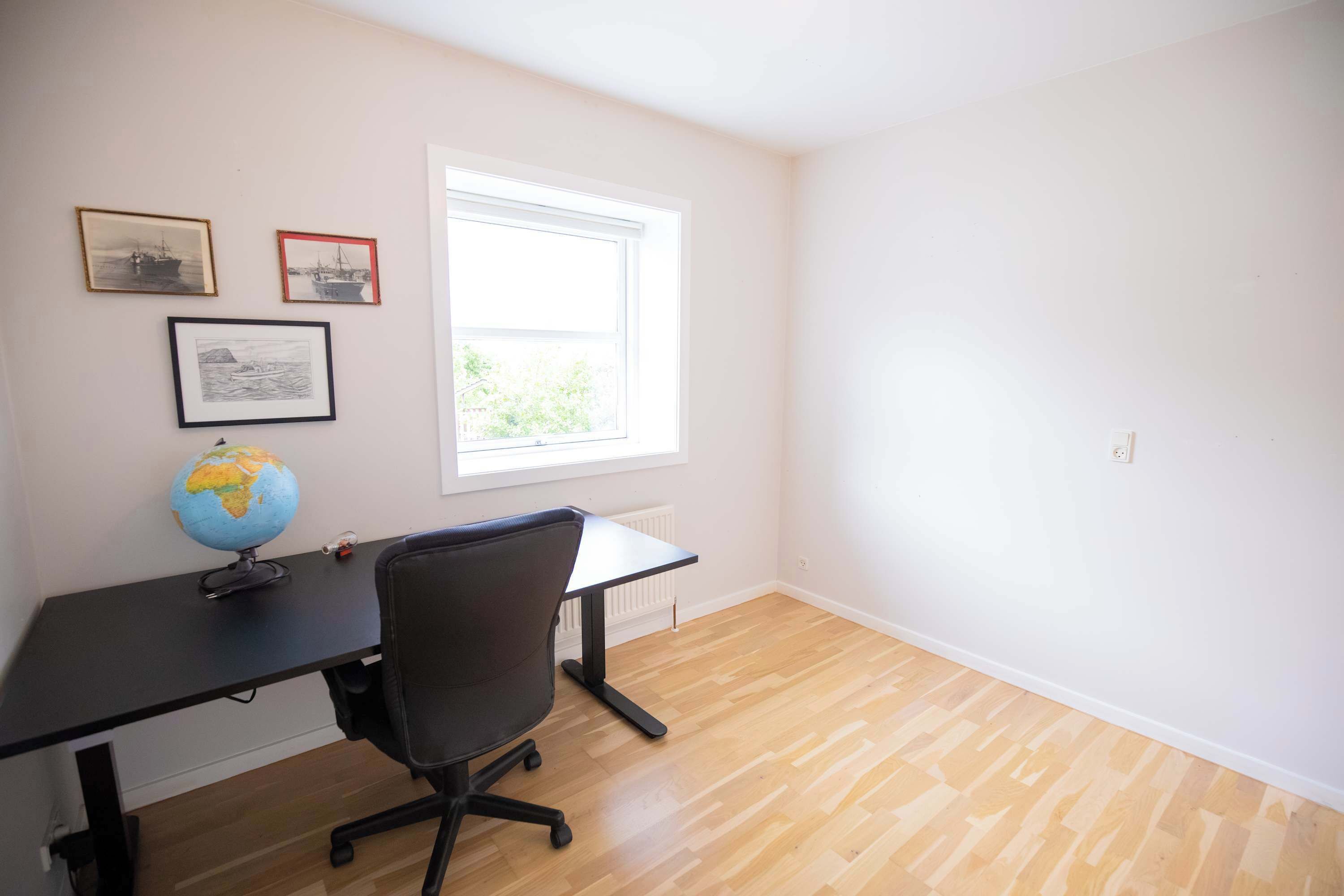 Une pièce avec un plancher en bois, un bureau noir surmonté d’un globe terrestre, une chaise et une fenêtre donnant sur un paysage verdoyant.