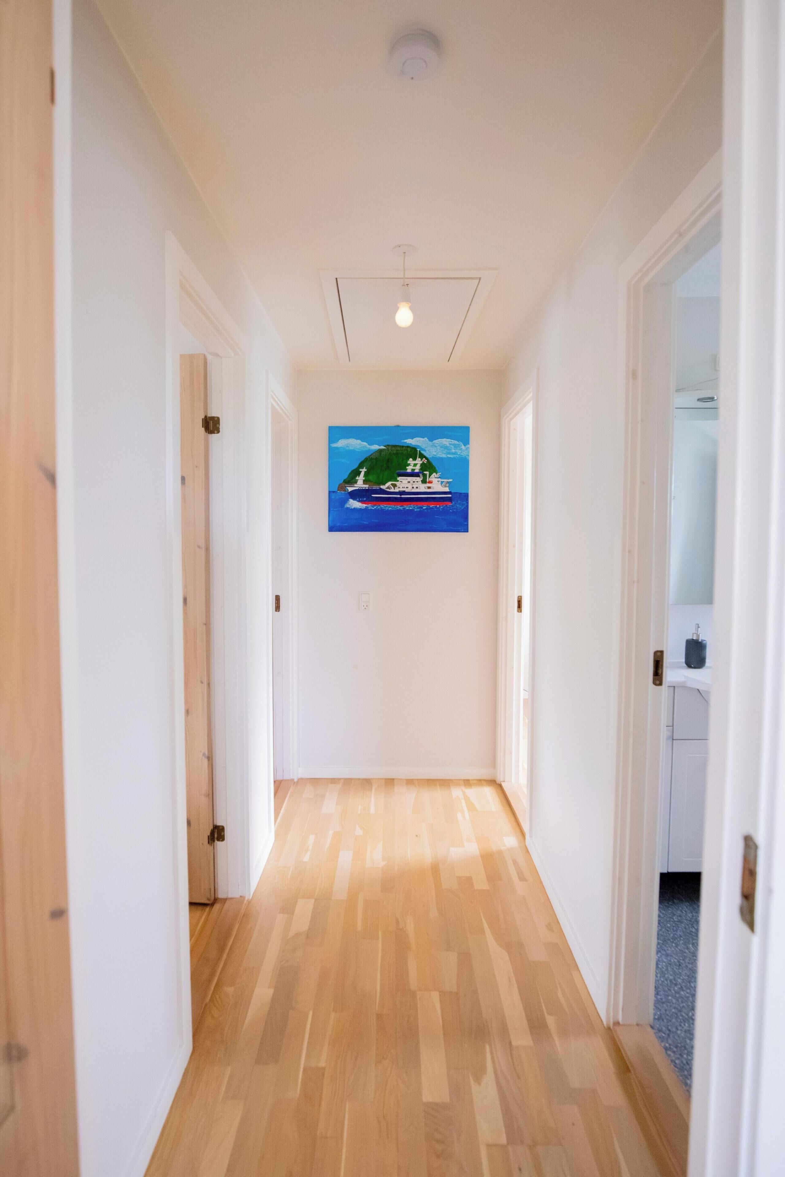 A hallway with wooden flooring leading to a room with a painting of a boat on a green island.