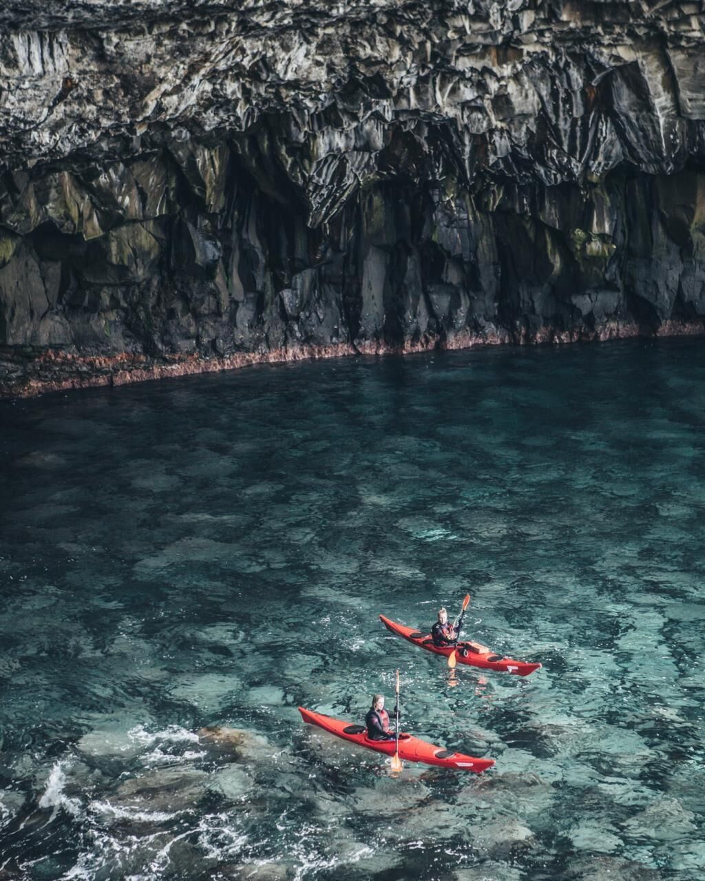 Kayakers in red kayaks on clear water with a rocky cliff in the background.