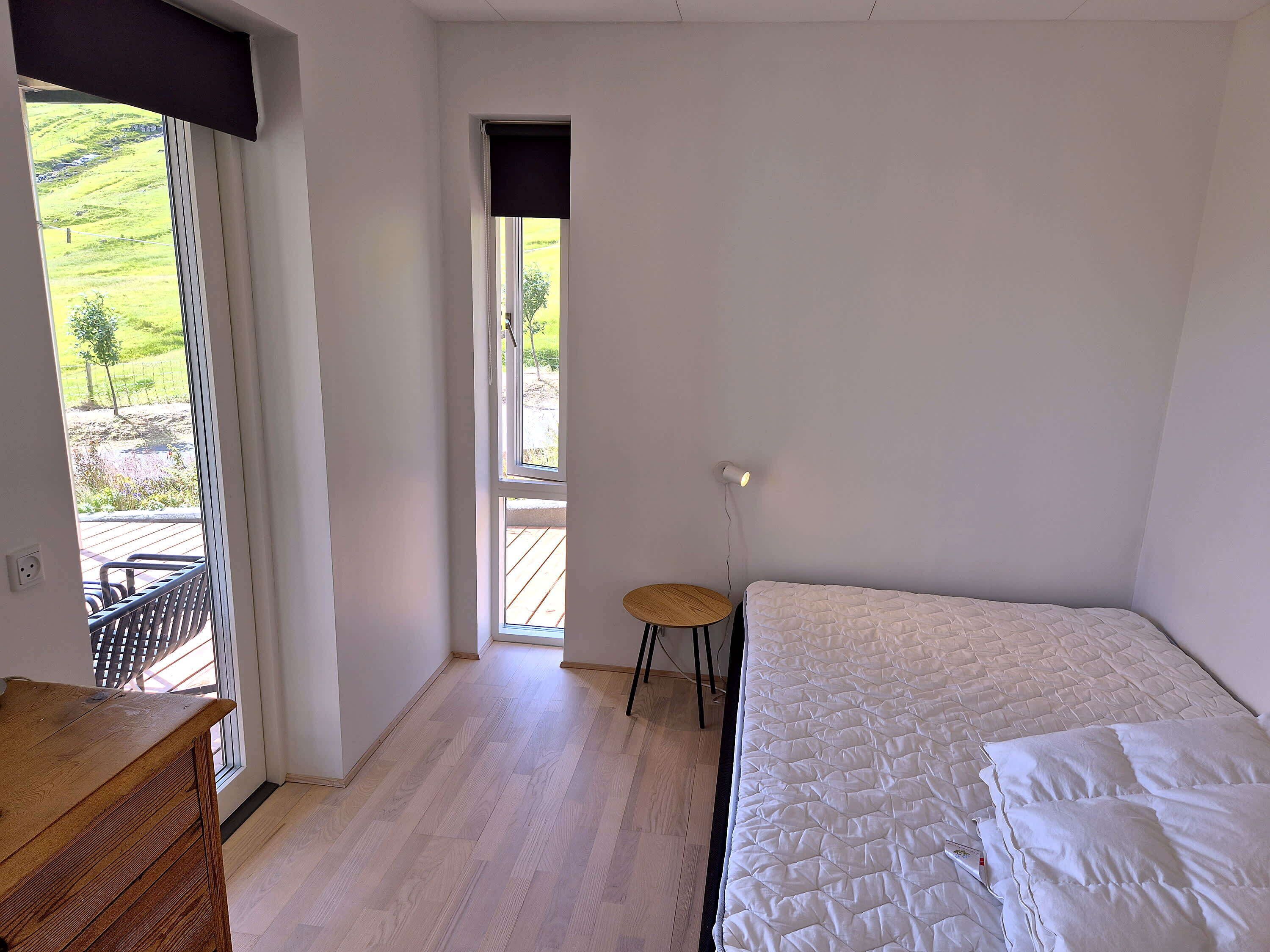 Een slaapkamer met een bed, een klein krukje en uitzicht op een tuin door een raam.