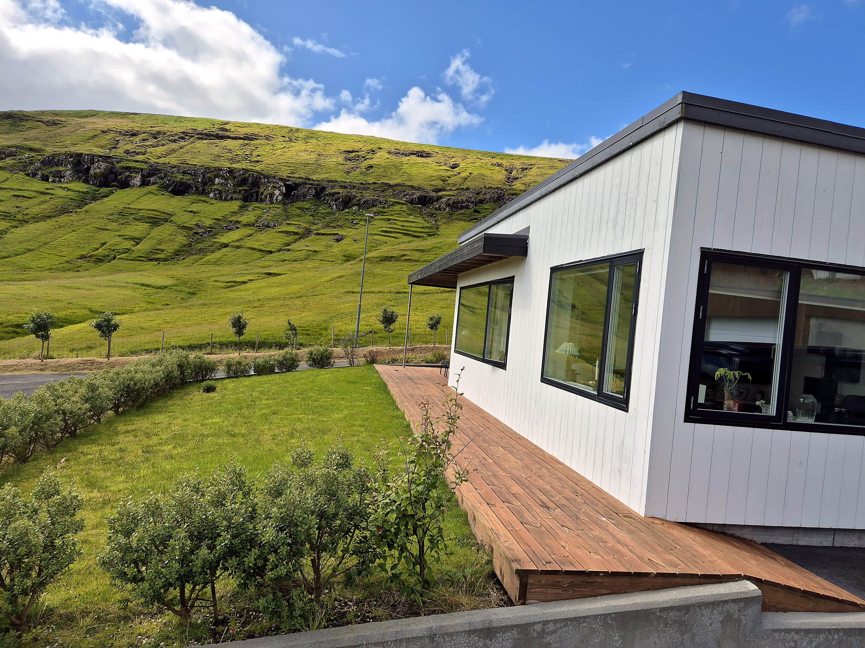 Una casa moderna con amplios ventanales, una terraza de madera y un paisaje con césped y colinas al fondo.