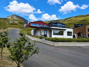 A modern residential area with contemporary houses, a curved road, and a mountain in the background. - Panoramic Fjord Views in Modern Kvivik House (Kvívík)