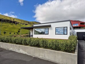 Une maison moderne à deux étages, dotée de grandes fenêtres, d’un toit rouge et offrant une vue sur une colline verdoyante.
