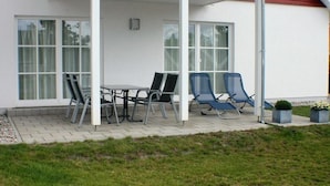 Ein überdachter Außenbereich mit Tisch und Stühlen, ein kleiner Garten und ein Gebäude mit großen Fenstern.