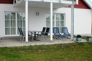 Ein überdachter Außenbereich mit Tisch und Stühlen, ein kleiner Garten und ein Gebäude mit großen Fenstern.
