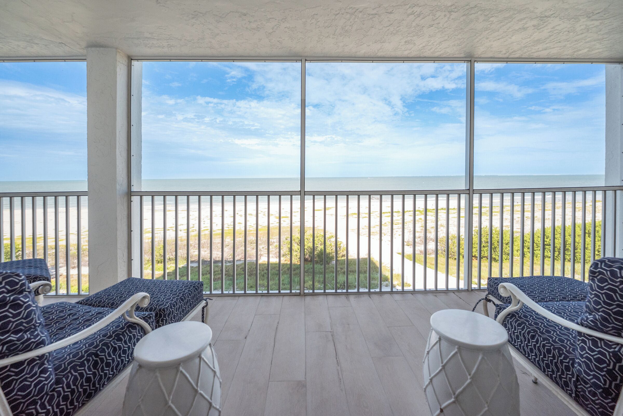 A balcony with two patterned chairs and a small table, overlooking a beach with a clear sky.