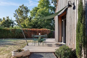 A patio with a table and chairs, a hanging light fixture, and a wooden deck leading to a building with a large window. - Holiday Home 'L'ancien Atelier' with Private Terrace, Wi-Fi and Air Conditioning (Muides-sur-Loire)