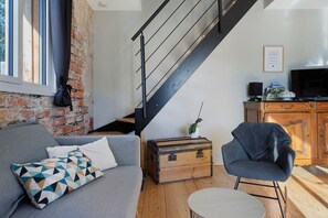 A modern living room with a grey sofa, a wooden chest, a small table, a chair, and a staircase. - Holiday Home 'L'ancien Atelier' with Private Terrace, Wi-Fi and Air Conditioning (Muides-sur-Loire)