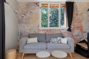 A room with a grey sofa, two round tables, a lamp, and a window with curtains. - Holiday Home 'L'ancien Atelier' with Private Terrace, Wi-Fi and Air Conditioning (Muides-sur-Loire)