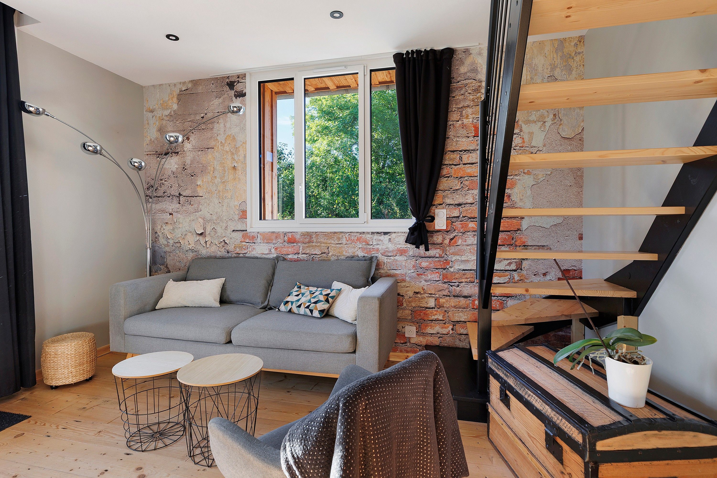 A cozy living room with a grey sofa, a small round table, and a wicker stool. A window with black curtains offers a view of greenery outside.