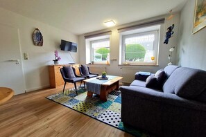 Una sala de estar con un sofá, dos sillones, una mesa de centro y un televisor. Hay ventanas con persianas y vistas a zonas verdes.