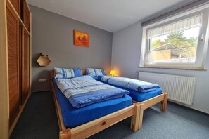 Un lit simple avec un cadre en bois et une literie bleue, une lampe de chevet, une fenêtre avec vue et une étagère fixée au mur.