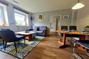 Una sala de estar moderna con un sofá, una mesa de centro y una mesa de comedor con sillas. Se ve una ventana con persianas, un radiador y una puerta al fondo.