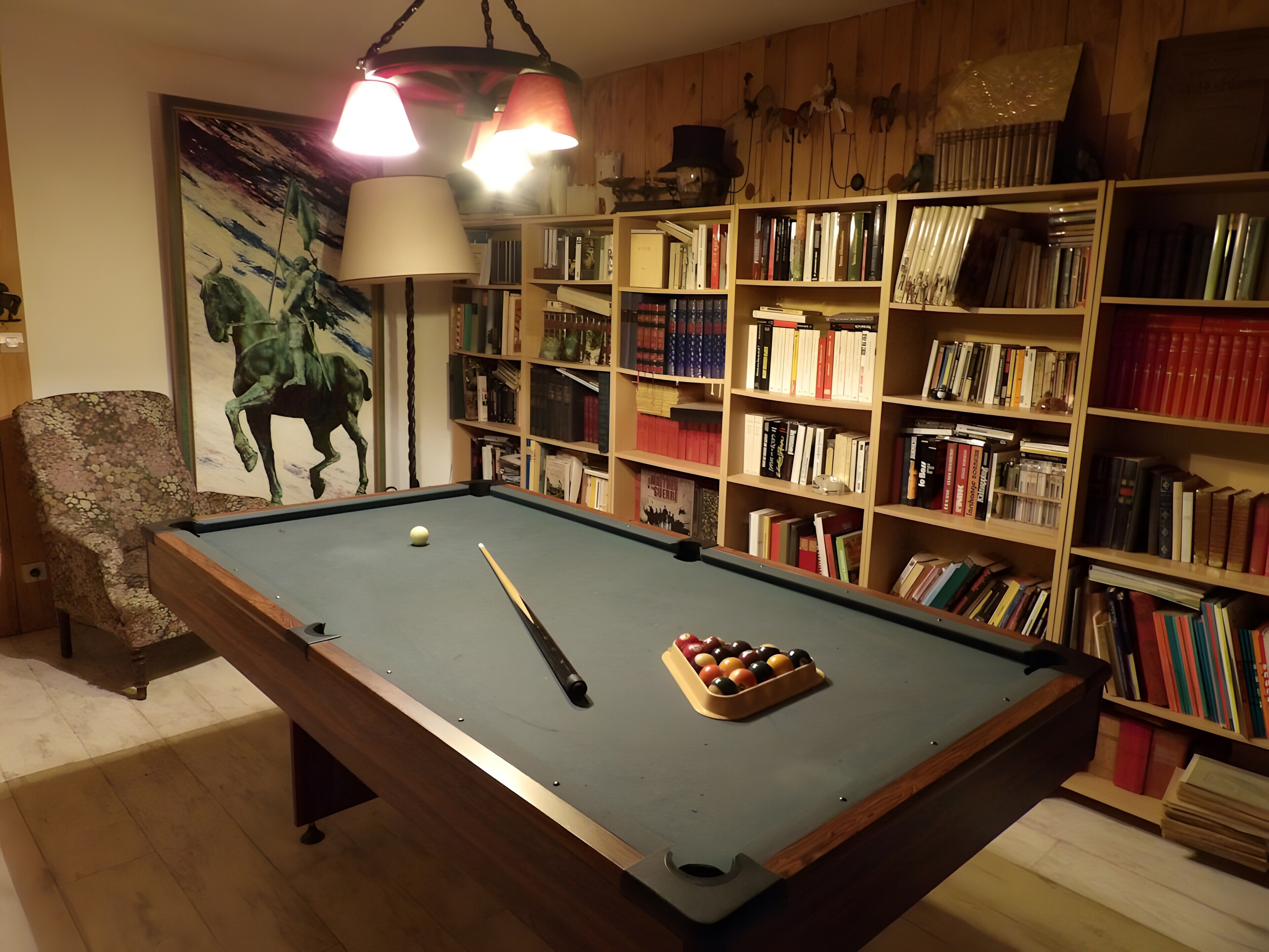 Una mesa de billar con taco y bolas, una silla y una estantería llena de libros.