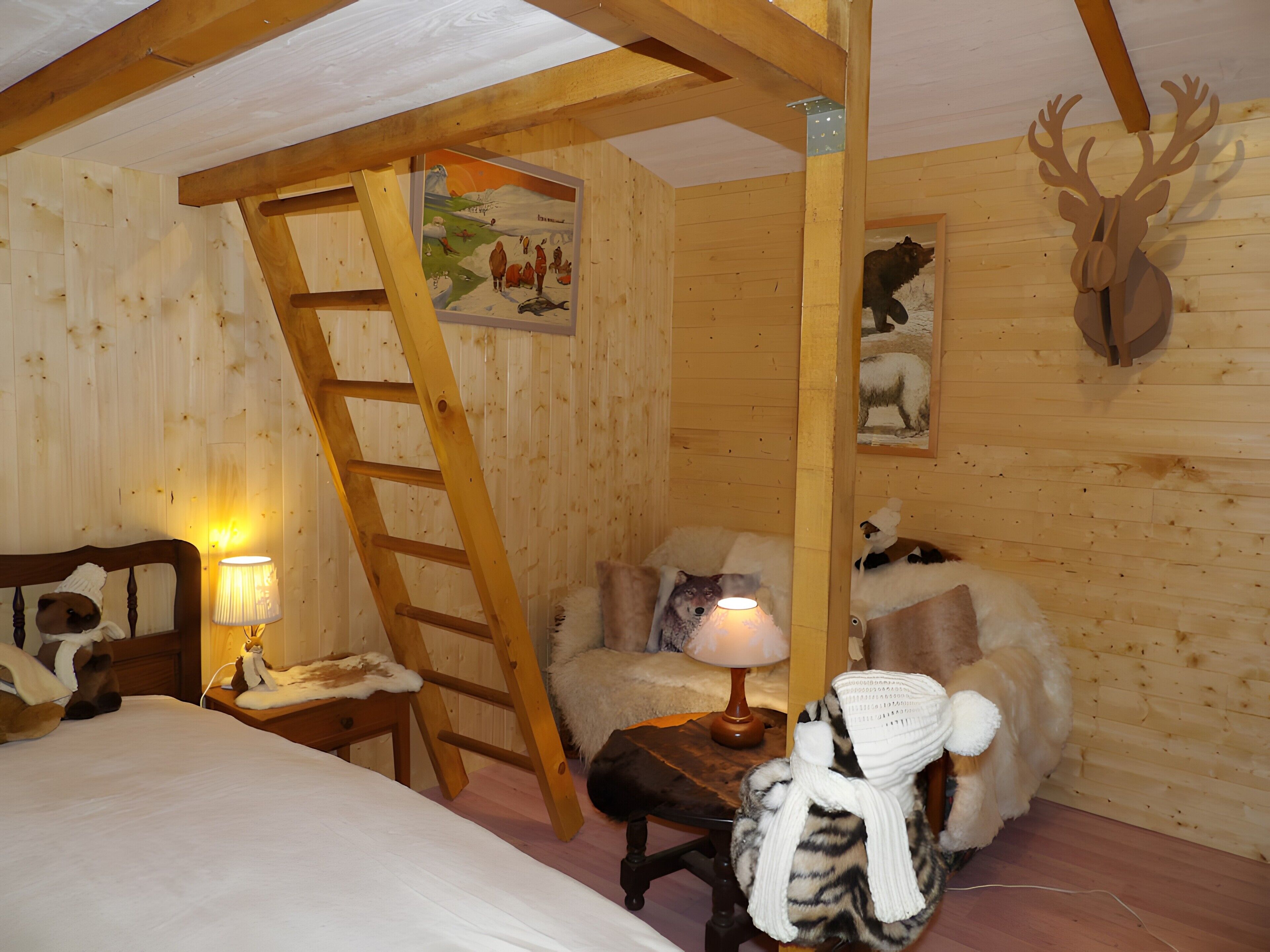 A cozy cabin room with a wooden ladder, a bed, a plush chair, and a wall-mounted deer head decoration.