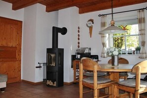 Een knus vertrek met een houtkachel, een eettafel met stoelen, een klok aan de muur en een raam met gordijnen.
