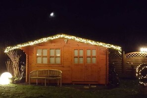 Een houten chalet met een bank en een fiets, omringd door een hek en verlicht met lichtslingers.