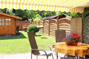Een terras buiten met een tafel, afgedekt met een gele tafelkleed, een stoel en een potplant met rode bloemen.