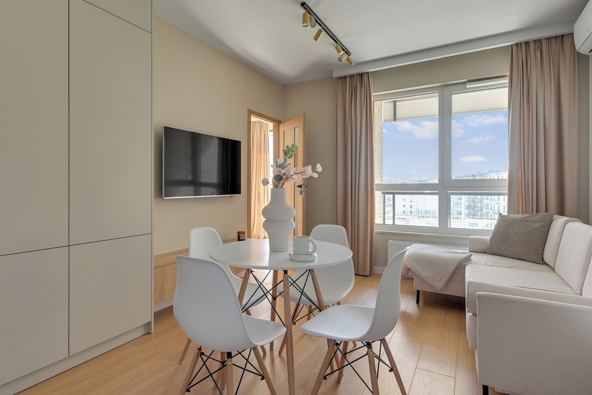 A modern living room with a flat-screen TV, a round dining table with chairs, a sofa, and a large window with curtains.
