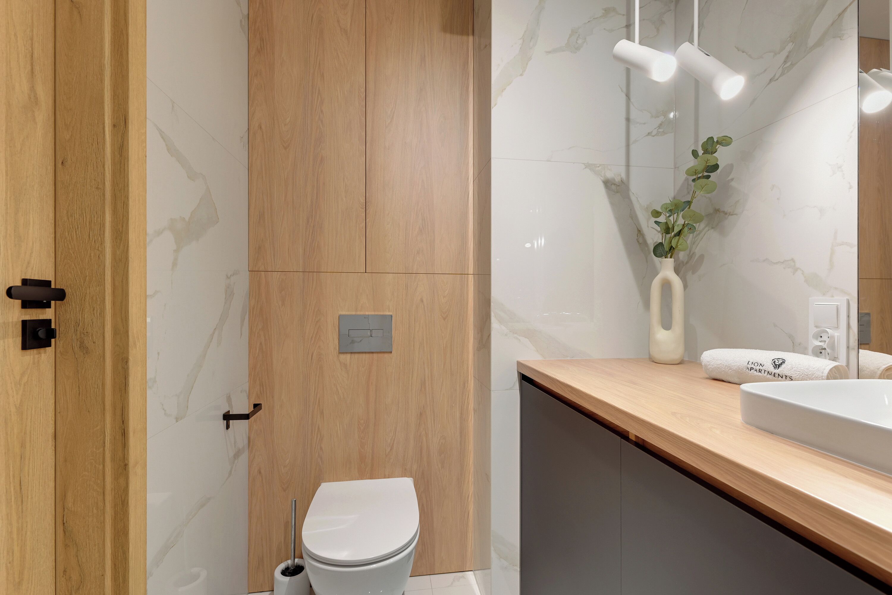 Une salle de bain moderne avec une porte en bois, des toilettes, un lavabo et un miroir agrémenté d’un vase contenant des plantes vertes.