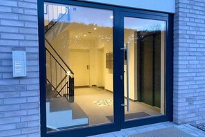A modern building entrance with a glass door, a visible staircase, and a small outdoor area. - Penthouse Passatstieg (Scharbeutz)