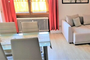 Un soggiorno con divano, tavolo da pranzo con sedie, termosifone e finestra con tende.