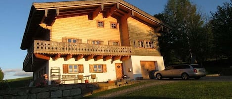 A wooden chalet with a balcony, stone wall, and a car parked in front.