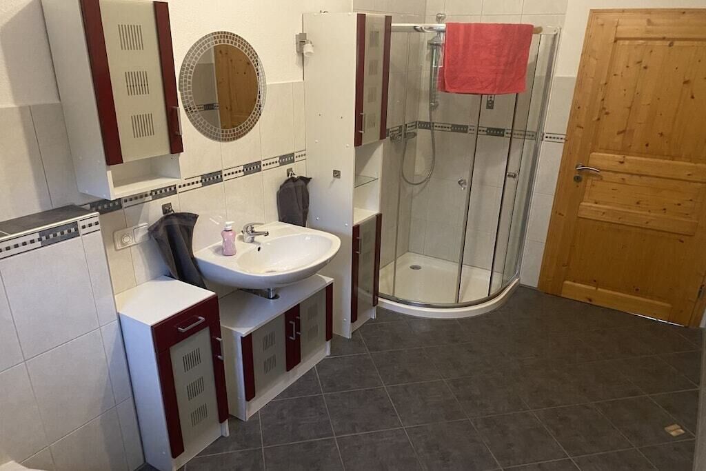 A bathroom with a white sink, a mirror, a shower enclosure, and a wooden door.
