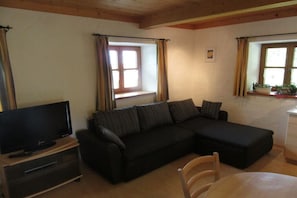 Ein Wohnzimmer mit einer schwarzen Couch, einem Fernseher auf einem Holzboden, einem Esstisch und Fenstern mit Vorhängen.
