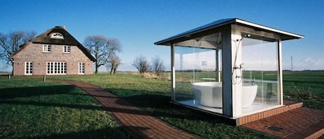A modern glass-enclosed structure with a toilet and shower, adjacent to a traditional brick house.