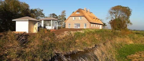 A traditional house with a thatched roof, a small adjacent structure, and a stream in the foreground.