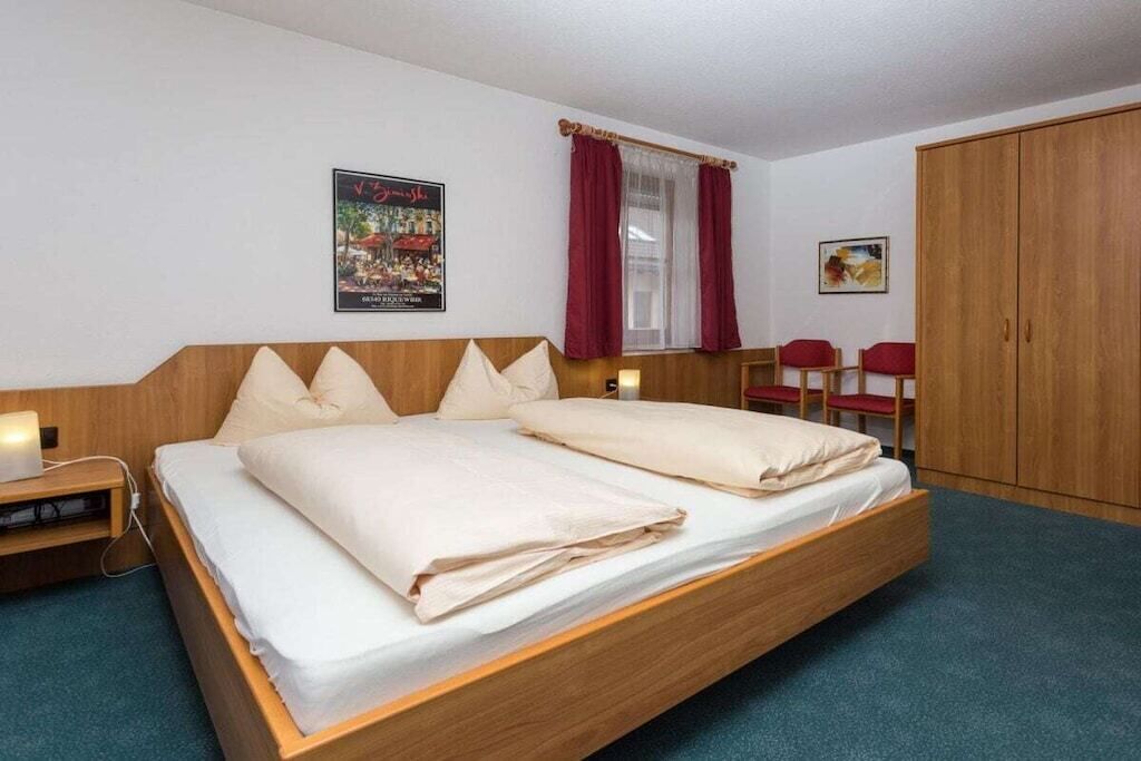 Uma cama de solteiro com estrutura de madeira e roupa de cama branca, uma mesa de cabeceira com abajur, um guarda-roupa e um quadro na parede.