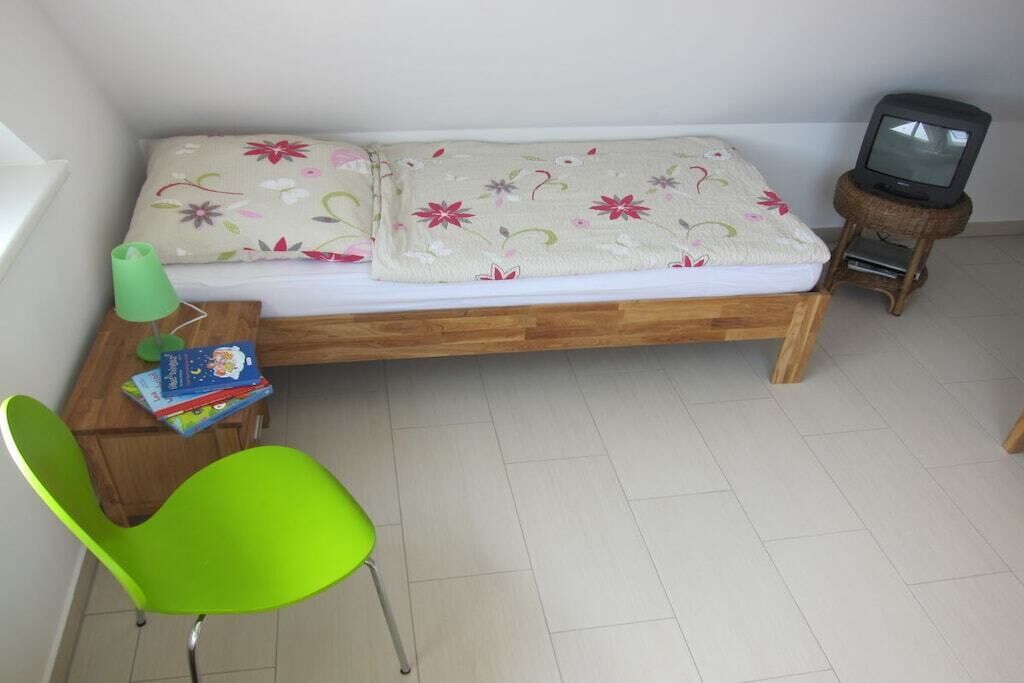 Una habitación pequeña con una cama individual, un televisor sobre un soporte, una silla verde y una mesita con libros.