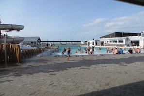 Een zwembadgedeelte met mensen die zwemmen en zonnebaden, een wachthoudershut en een gebouw met grote ramen.