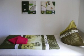 Een bed met een kussen met een groen-wit patroon, een rode handdoek en twee abstracte schilderijen aan de muur.