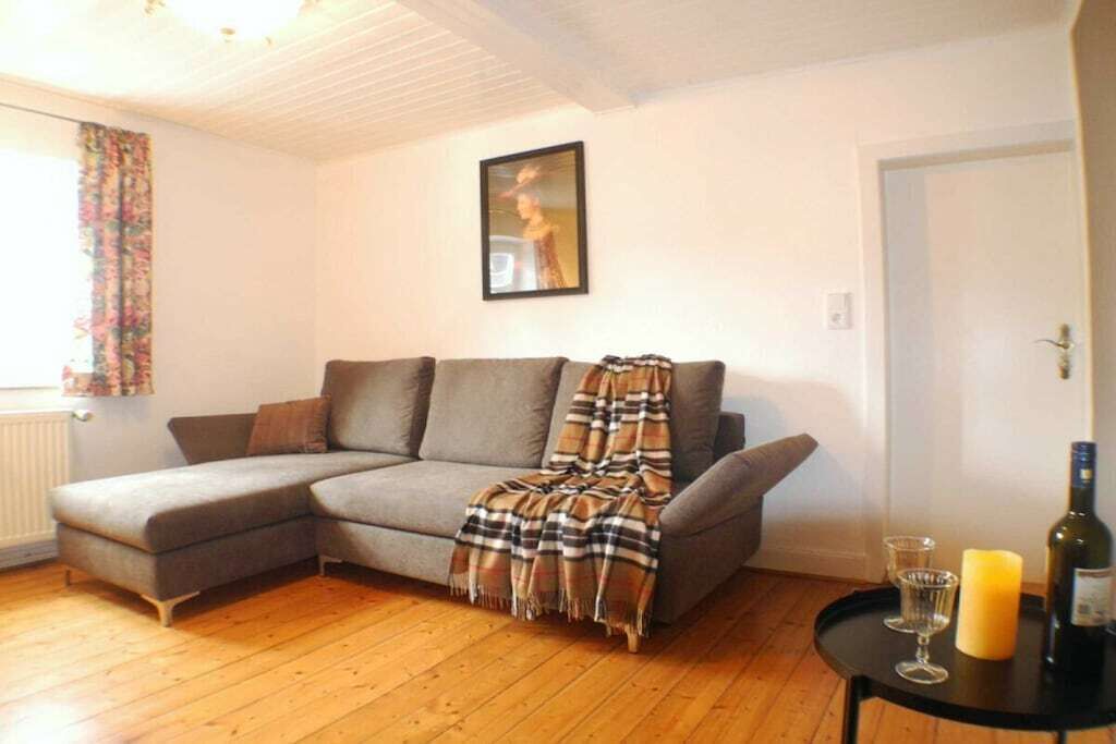 Ein Wohnzimmer mit einem grauen Ecksofa, einer karierten Decke, einem gerahmten Bild an der Wand und einem kleinen Tisch mit Kerze und Flasche.