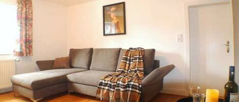 Ein Wohnzimmer mit einem grauen Ecksofa, einer karierten Decke, einem gerahmten Bild an der Wand und einem kleinen Tisch mit Kerze und Flasche.