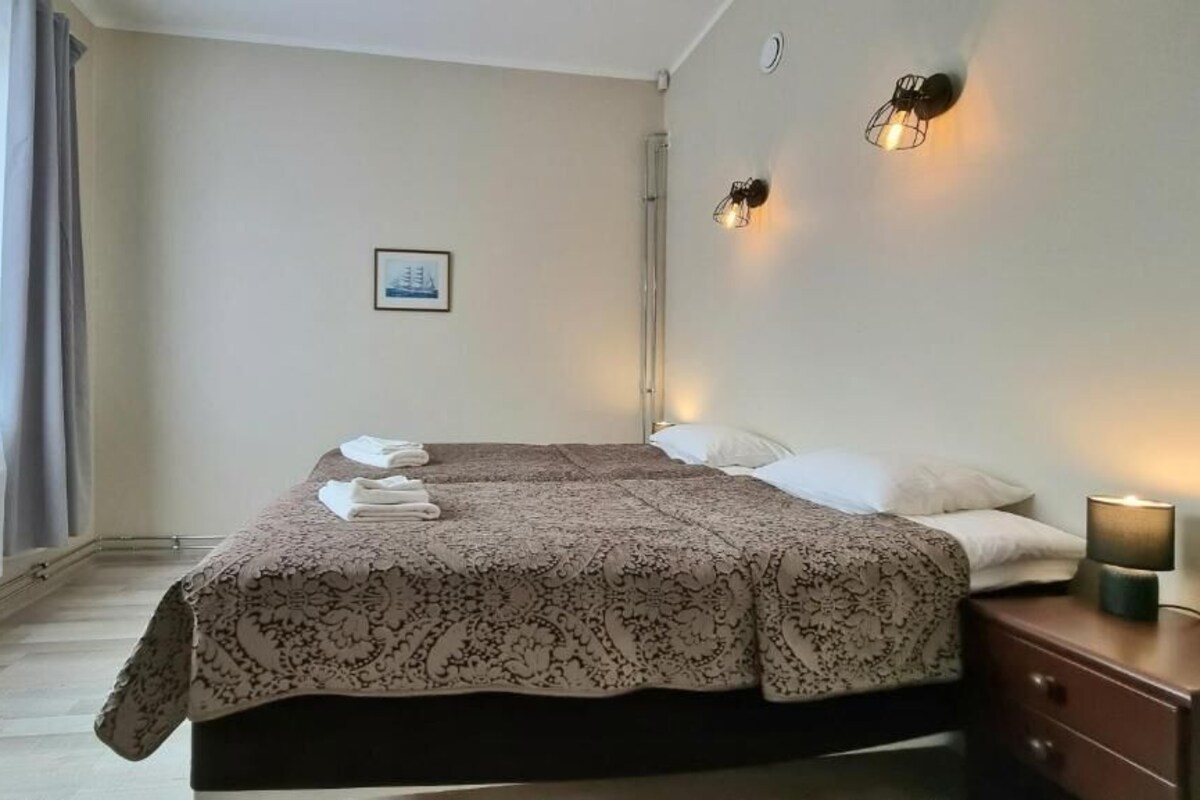 Una habitación de hotel con cama, mesita de noche, lámpara y ventana con cortinas.
