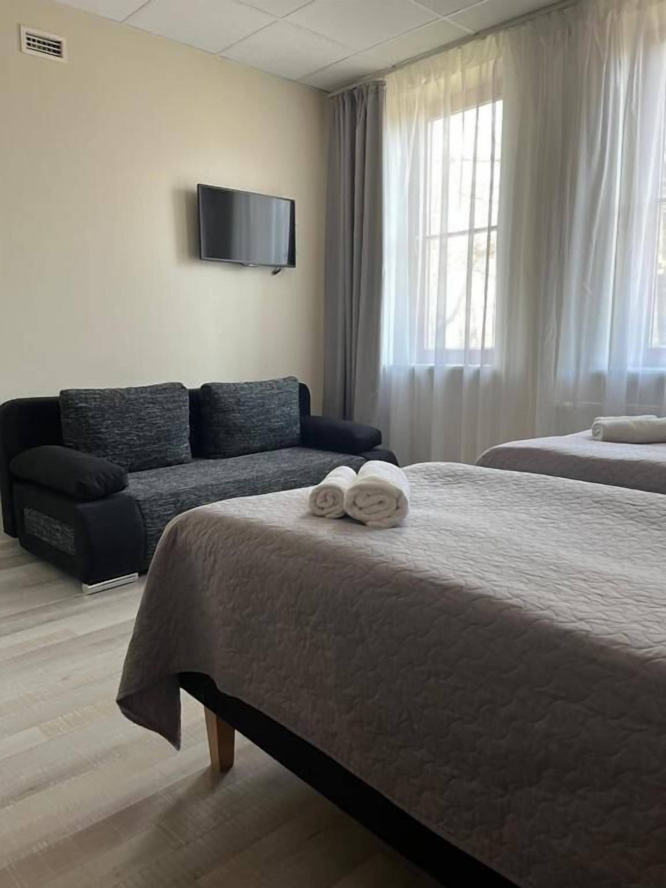 A hotel room with a bed, a sofa, a flat-screen TV, and large windows with curtains.
