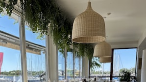 A restaurant with wooden tables and chairs, large windows with a marina view, and hanging wicker lamps.