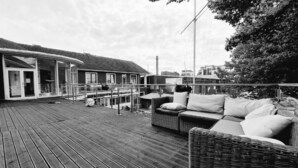 Una terraza de madera con mobiliario exterior, un edificio con cerramiento de vidrio y un cielo despejado.