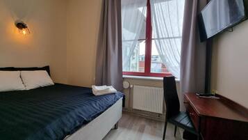 Et hotelværelse med seng, skrivebord, stol, radiator og et fjernsyn ophængt på væggen.