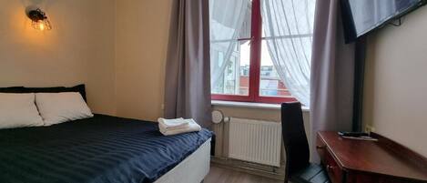 A hotel room with a bed, a desk, a chair, a radiator, and a TV mounted on the wall.