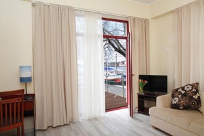 Una habitación con una silla de madera, un televisor sobre un mueble bajo, un sofá con un cojín estampado y una puerta que da a un espacio exterior con vistas a barcos.