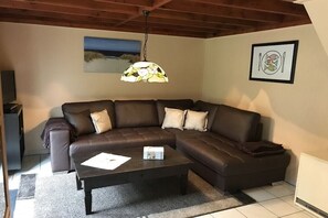 Een woonkamer met een hoekbank, een salontafel, een hanglamp, een lijst met een foto aan de muur en een radiator.