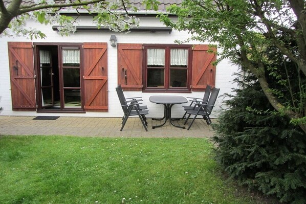 Een wit gebouw met houten luiken, een tafel met stoelen en een gazon ervoor.