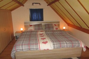 Een slaapkamer met een houten bed, een hoofdbord, twee bedlampjes, een raam met gordijnen en een houten plafond.