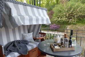 A balcony with a striped awning, a cushioned seating area with a pillow, a small table with glasses and condiments, and a view of greenery. - "Baltic Sea Love" Villa Lena Wilhelmstrasse (Sellin)
